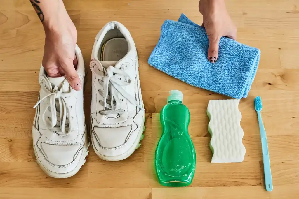 A top view of a person holding a pair of whitw sneakers and a dish soap bottle and a sponge and and toothbrush with a blue cloth